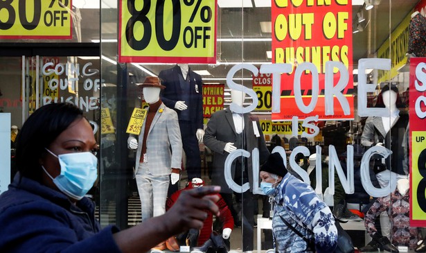 A woman wearing a mask stands in front of a store going out of business, with "STORE IS CLOSING" written on the window and 80% off signs