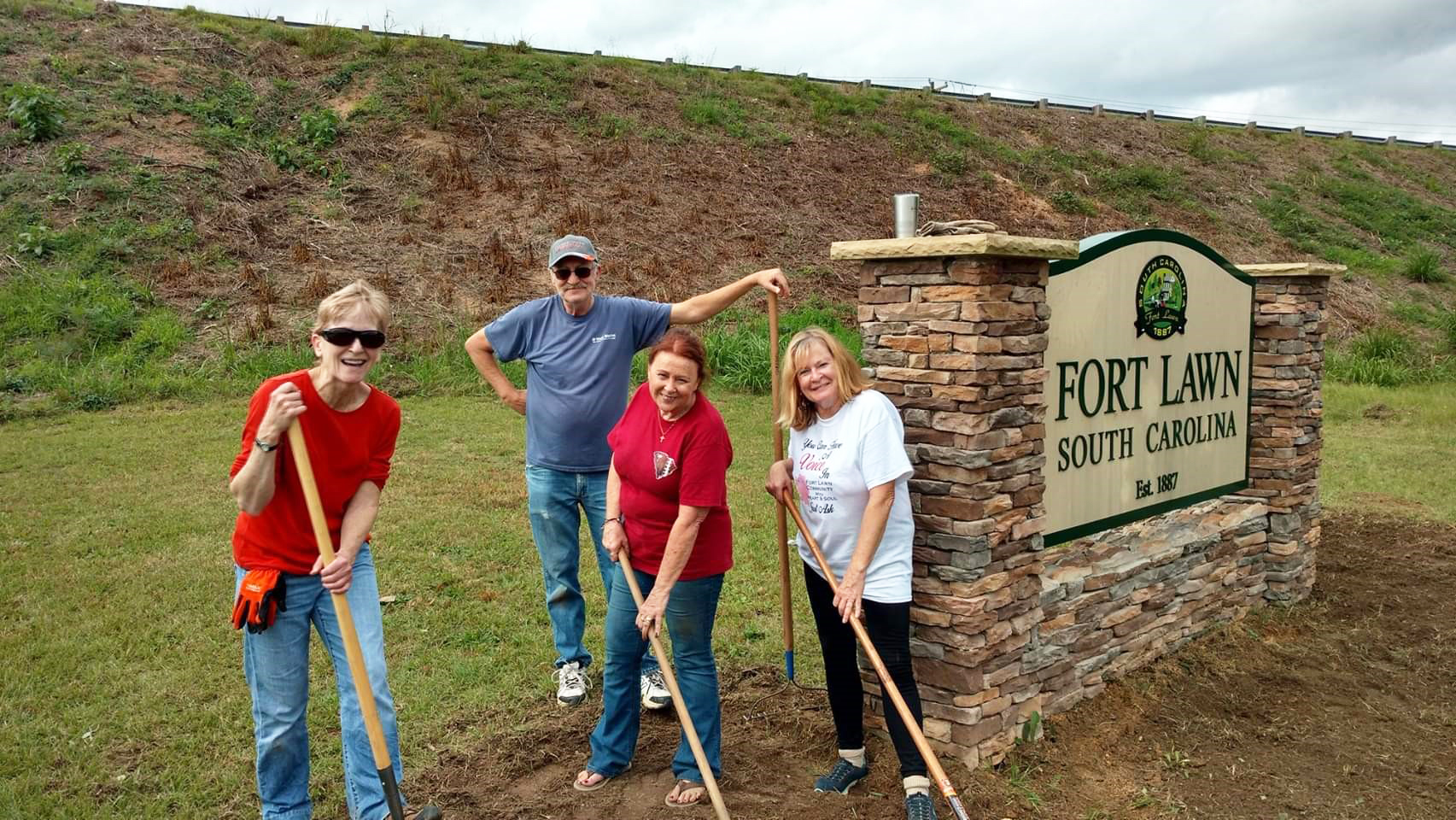 Community members holding rakes and shovels pose in front of Fort Lawn, South Carolina town sign.