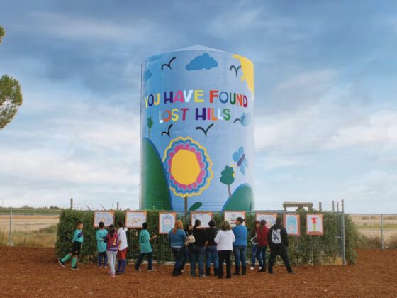 Image of children playing around a water tower, with bright paintings on it and the message "You Have Found Lost Hills"